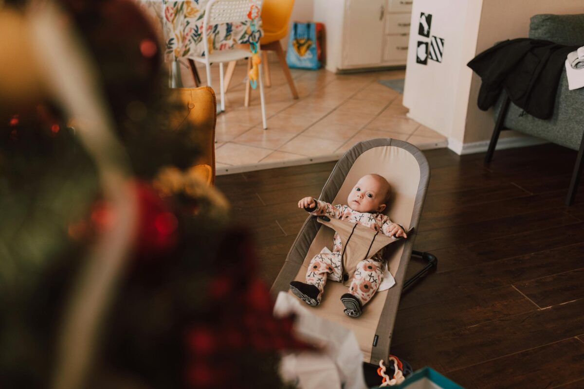 A cute baby in a baby bouncer seat gazes at a Christmas tree in a cozy living room setting.