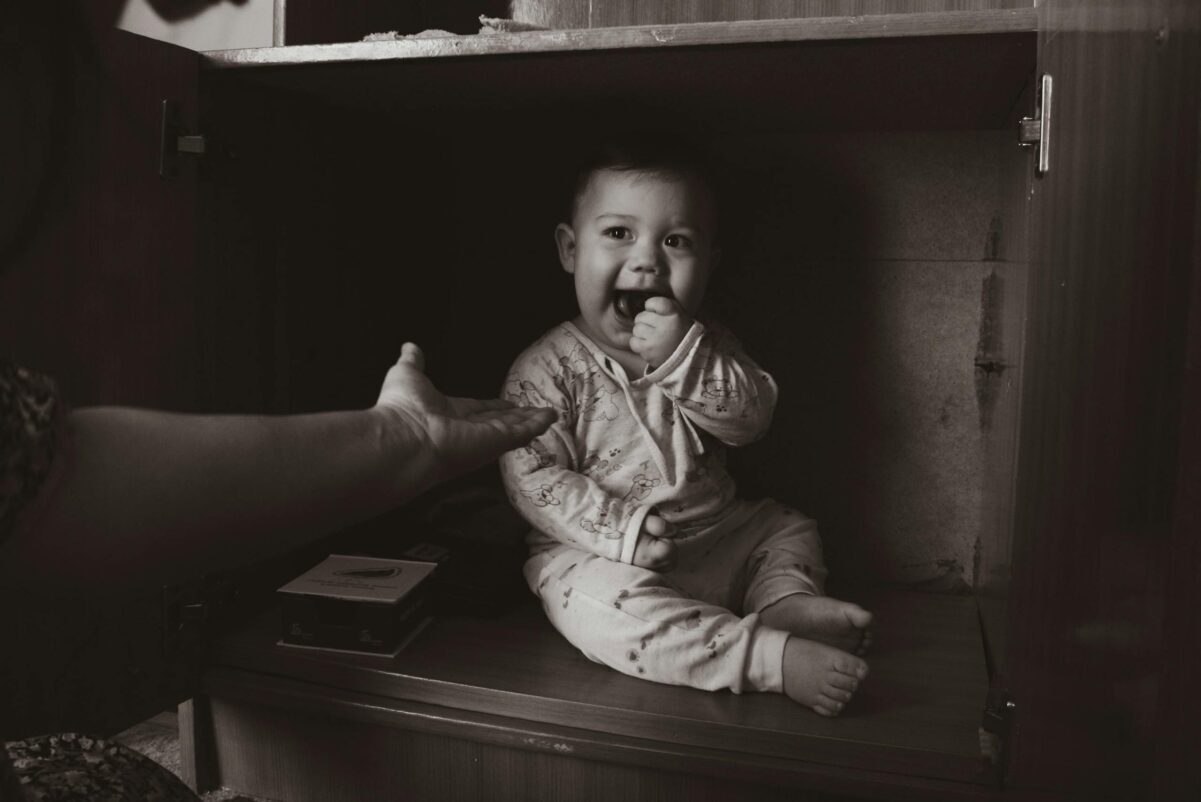 babyproofing: Charming grayscale photo of a baby sitting inside a cabinet, reaching out to a hand.