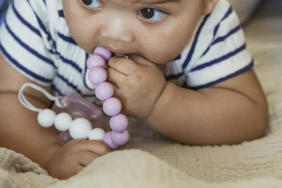 Cute little African American baby with curly hair lying on sofa while playing with beads in room on blurred background teething