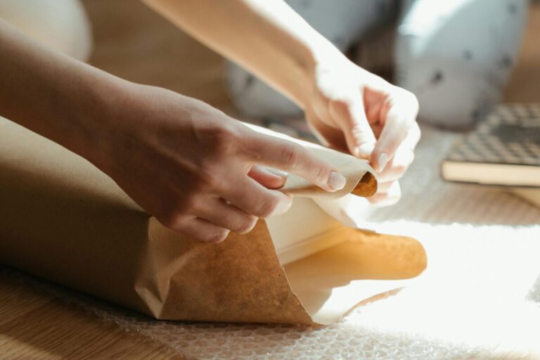 Hands wrapping an item in brown paper with sunlight streaming in, highlighting the texture and details. new mom gift