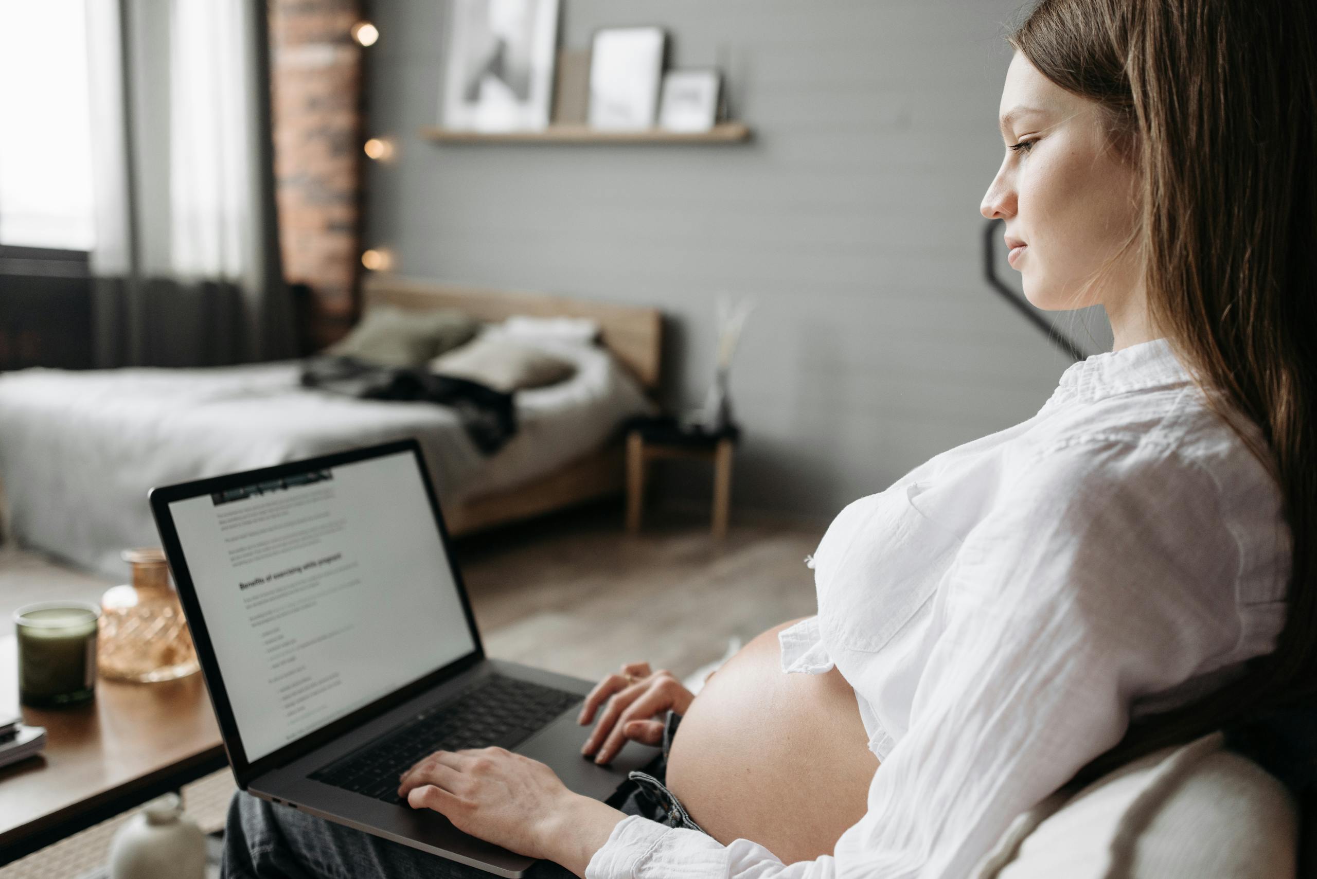 Pregnant woman working on a baby registry on a laptop in a cozy, light-filled bedroom. Side view of her baby bump.
