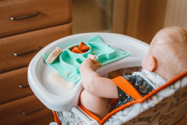 A cute baby enjoying baby-led weaning snack time in a high chair with colorful food tray indoors.