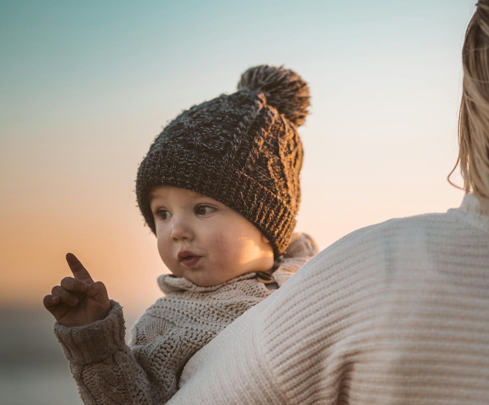 cold weather baby essentials: A mother carries her bundled-up toddler at a beach during a picturesque winter sunset.