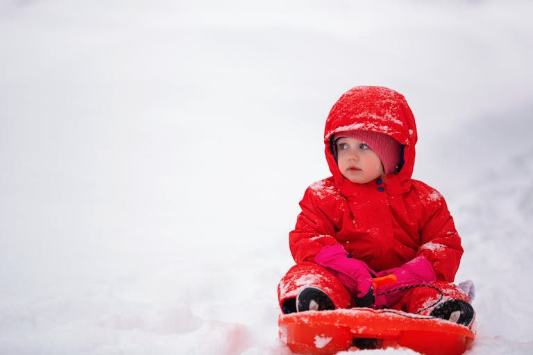 Cold weather baby essentials: Cute baby in bright red snowsuit playing in the snow on a cold winter day.